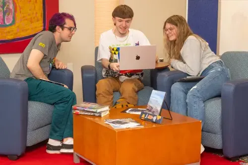 A group of three students sitting on arm chairs review material on the center student's laptop